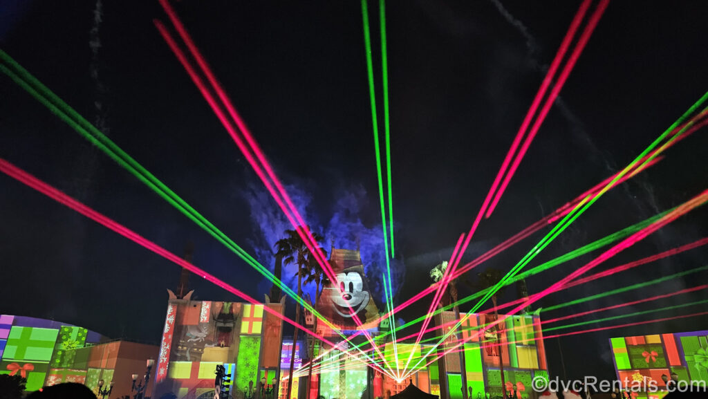 Red and green lasers fill the sky above the Chinese Theater during Jingle Bell, Jingle BAM! The theater is lit up with red and green projections that look like gift boxes, as well as Mickey Mouse at the top of the building.