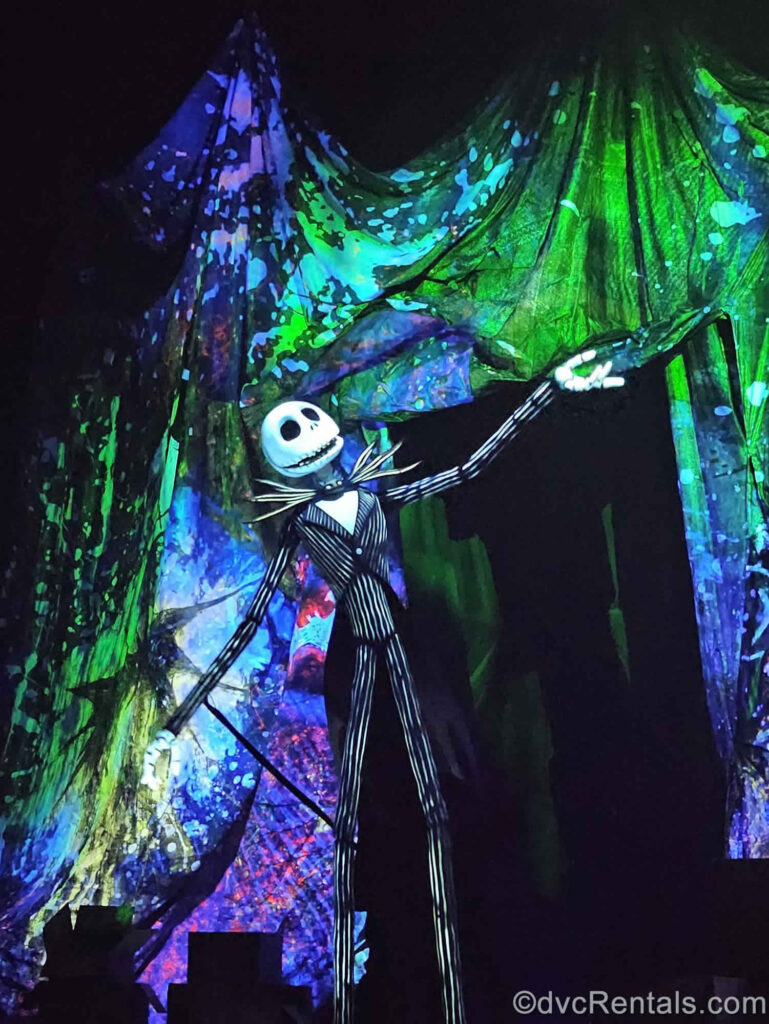 A black and white life-size Jack Skellington puppet performs on stage in front of purple, blue, and green curtains. Jack’s arms are outstretched, and he is looking upwards to the right.