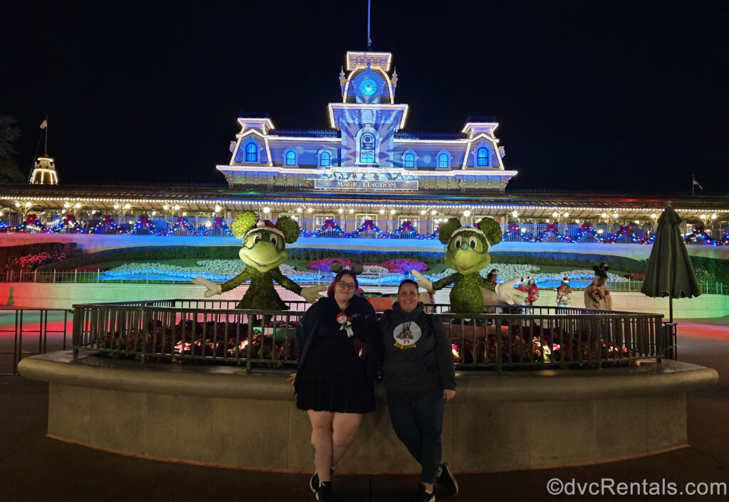 Team members Kristen and Stacy pose in front of Mickey and Minnie Mouse topiaries and the Magic Kingdom Train Station, which is lit up blue, green, red for Christmas.