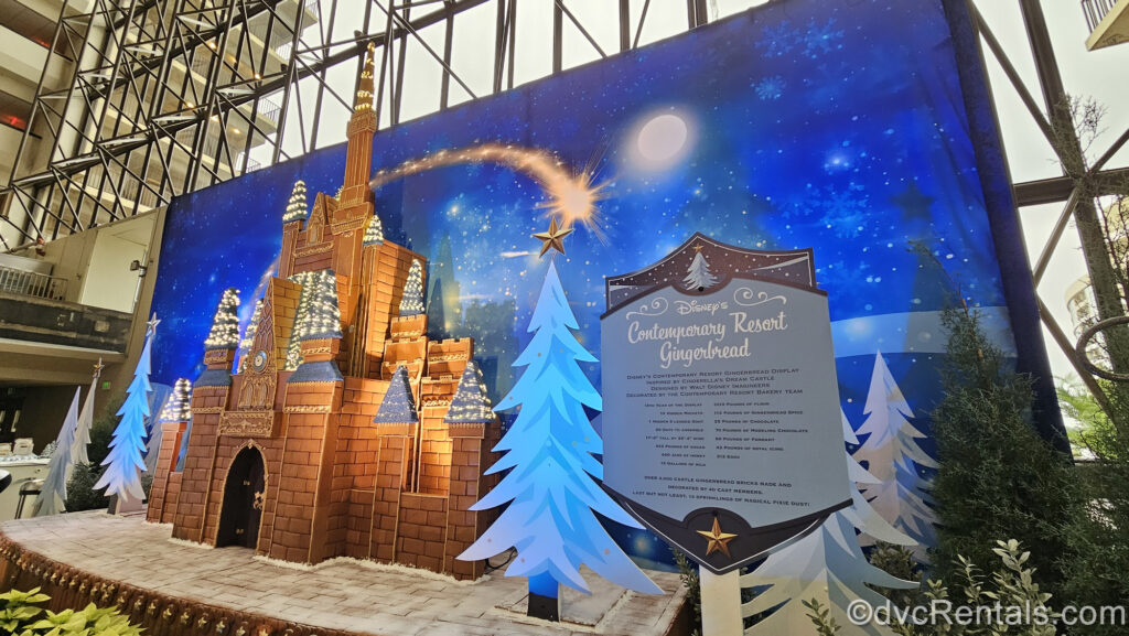 A large gingerbread version of Cinderella Castle is displayed in front of a blue, wintery background at Disney’s Contemporary Resort. To the right of the gingerbread castle is a blue sign listing the ingredients used to make the gingerbread.