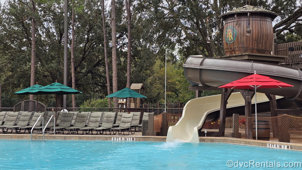 The brown and beige twisting tube waterslide at pool. To the left of the slide there are many green loungers, and a wooden water tower sits atop the waterslide.