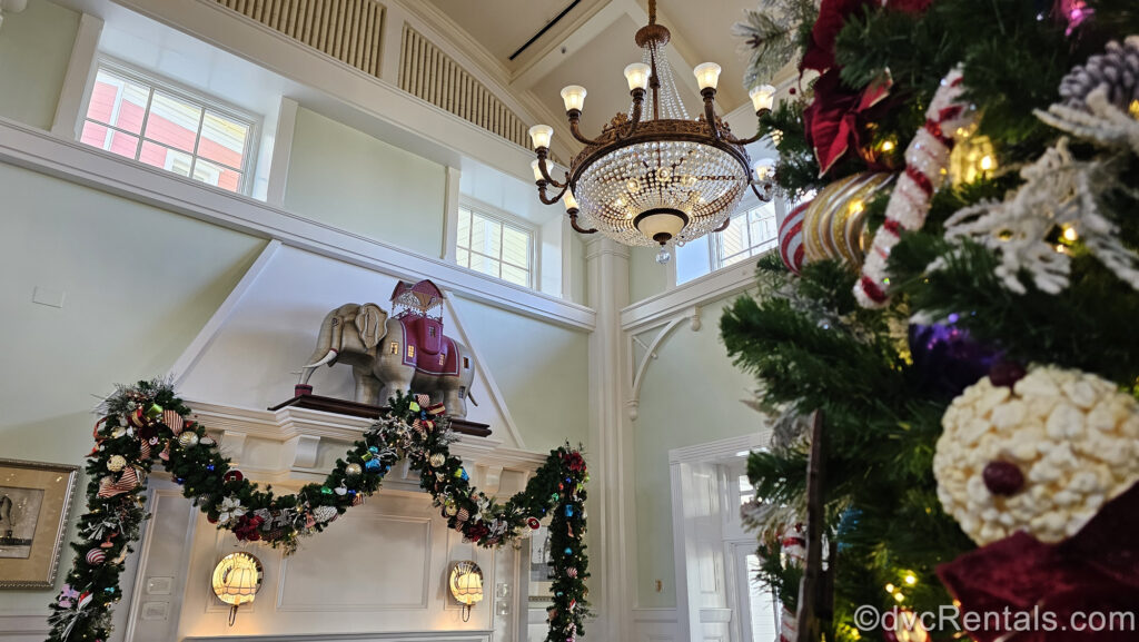 Green garland and a green Christmas tree both with multicolored ornaments decorate the pale green lobby of Disney’s BoardWalk Villas. Over the garland there is a circus elephant figure and a crystal chandelier hangs overhead.