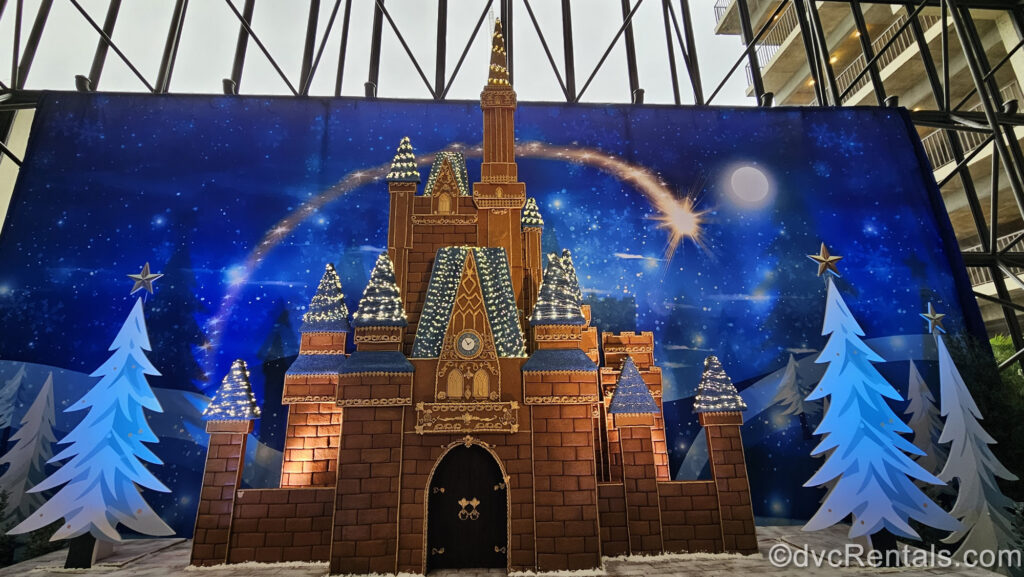 A gingerbread creation made to look like Cinderella Castle at Disney’s Contemporary Resort. There are blue and white wooden trees to either side of the castle, and a blue starry night background with a shooting star sits behind it.