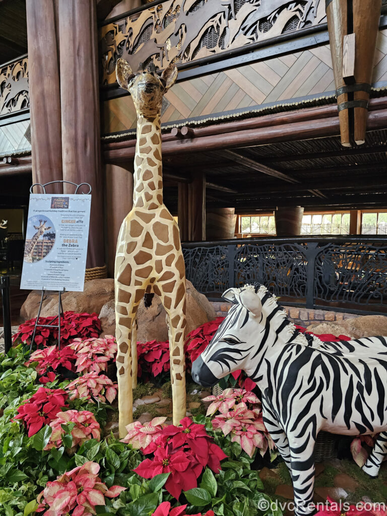 Gingerbread creations made to look like a baby giraffe and baby zebra are on display at Disney’s Animal Kingdom Villas Jambo House. They are surrounded by red and white poinsettia plants.