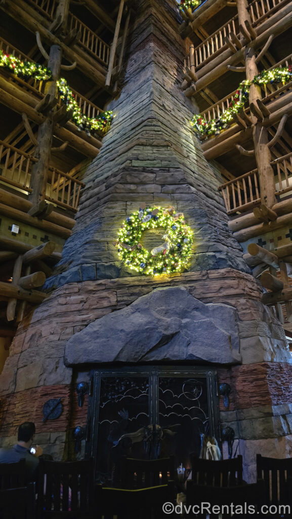 Green garlands and a wreath decorated with white lights and rustic ornaments decorate the lobby of Disney’s Wilderness Lodge. The wreath is set upon the large stone fireplace with rocking chairs in front of it.