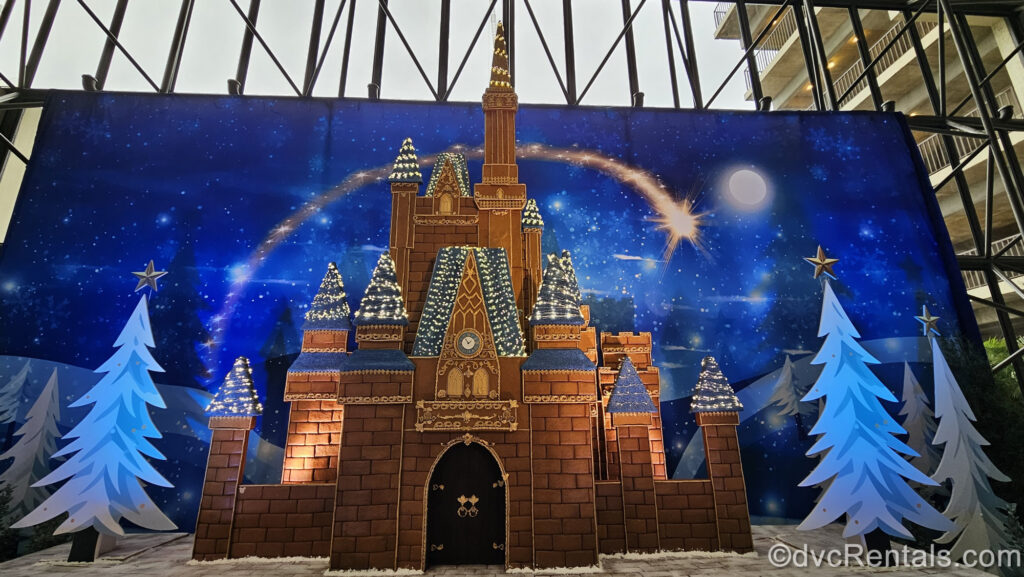 a large-scale replica of Cinderella Castle made of gingerbread is displayed at Disney’s Contemporary Resort in front of a blue backdrop meant to look like a wintery night. The castle peaks are royal blue with tiny white lights, and to each side of the castle there are white and blue tree cutouts.