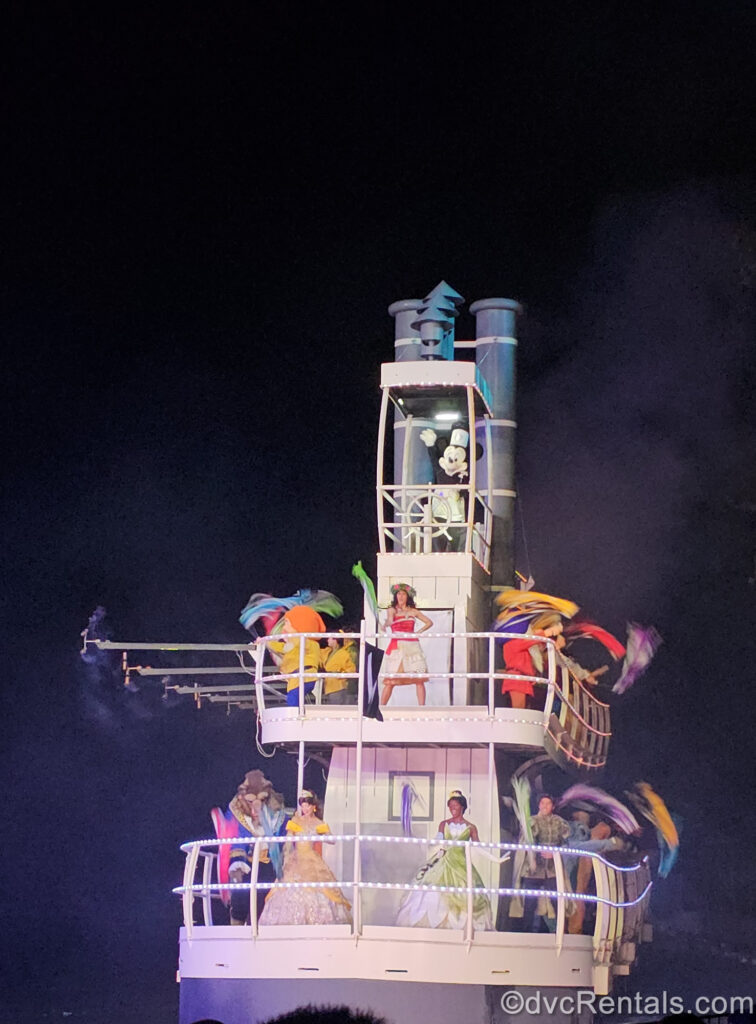 Characters including Steamboat Willie, Moana, Beast, Belle, Tiana, Naveen, and the 7 Dwarfs wave ribbons on a white steamboat float during Fantasmic!