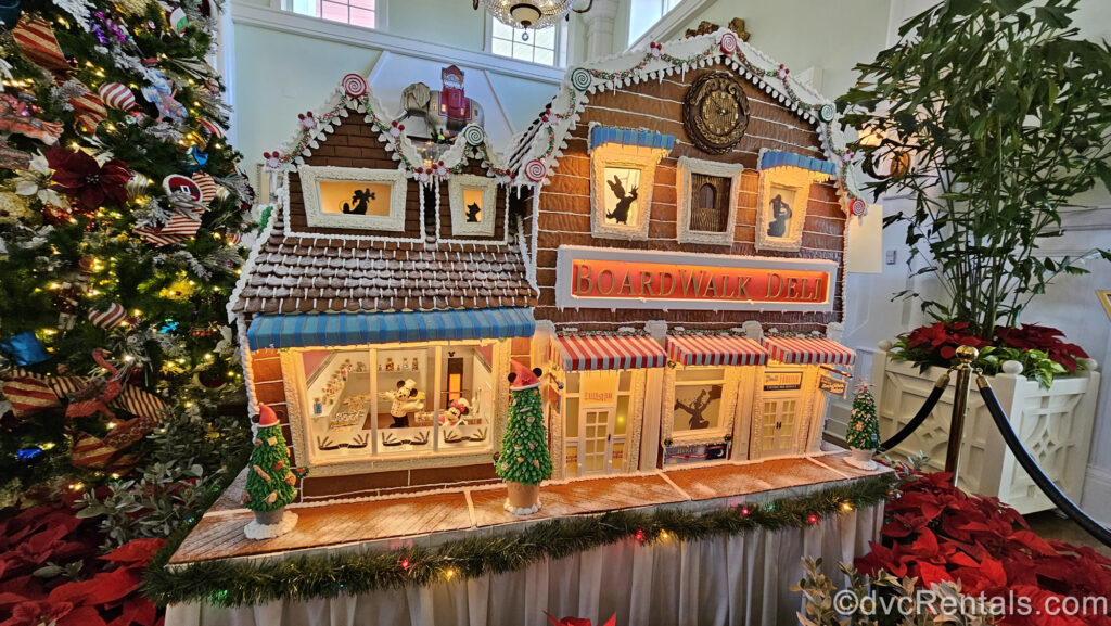 A gingerbread replica of the BoardWalk Deli is displayed at Disney’s BoardWalk Villas. Disney characters decorate the gingerbread display, and a tall Christmas tree decorated with multicolored ornaments and white lights is visible to the left of the display.