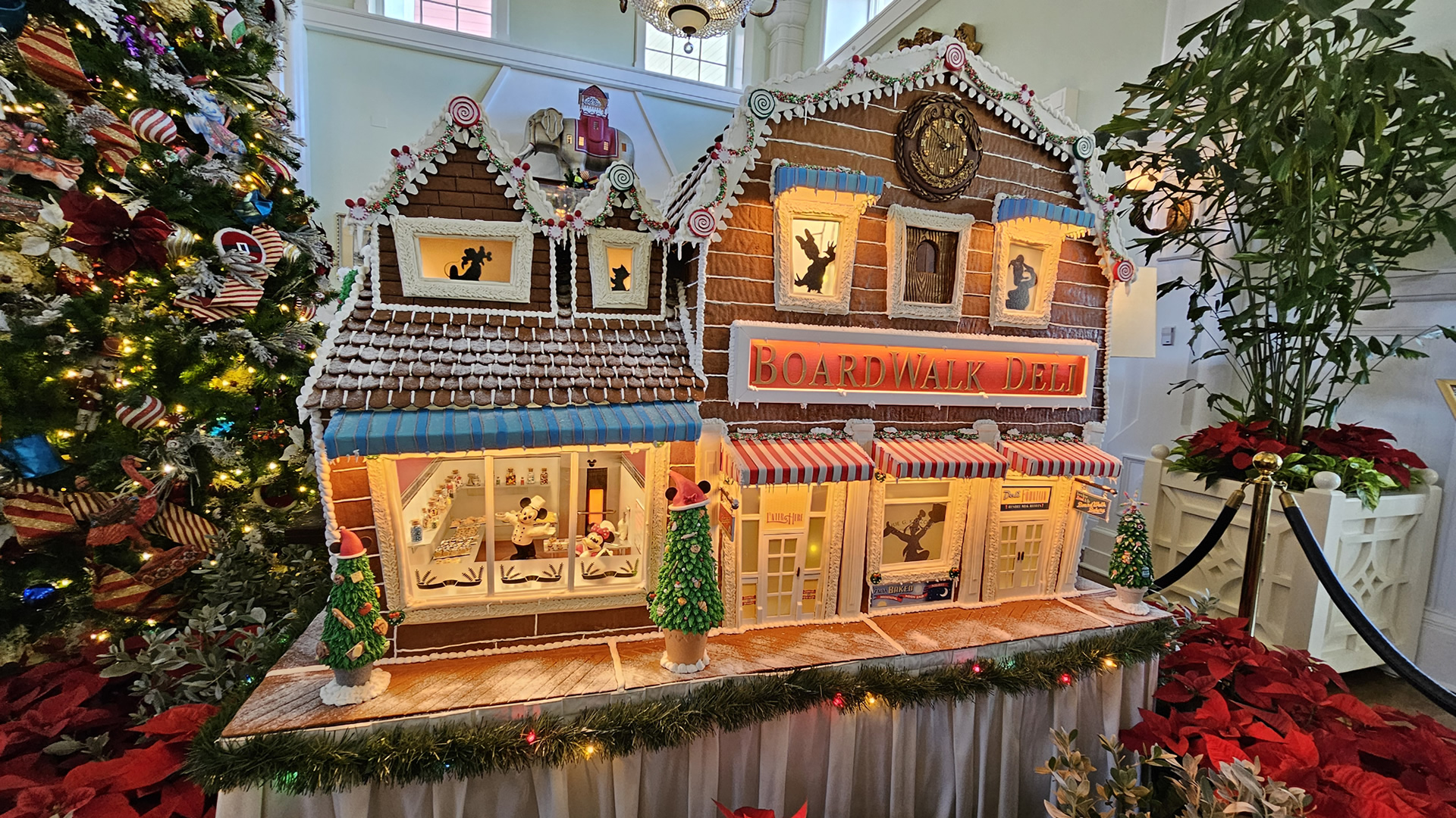 A gingerbread replica of the BoardWalk Deli is displayed at Disney’s BoardWalk Villas. Disney characters decorate the gingerbread display, and a tall Christmas tree decorated with multicolored ornaments and white lights is visible to the left of the display.