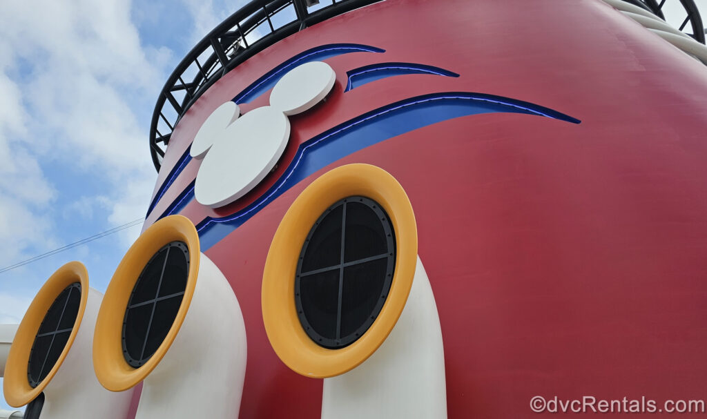 The red funnel of a Disney cruise ship is seen under a blue sky. The funnel features the blue and white Disney Cruise Line logo and yellow and white horn speakers.