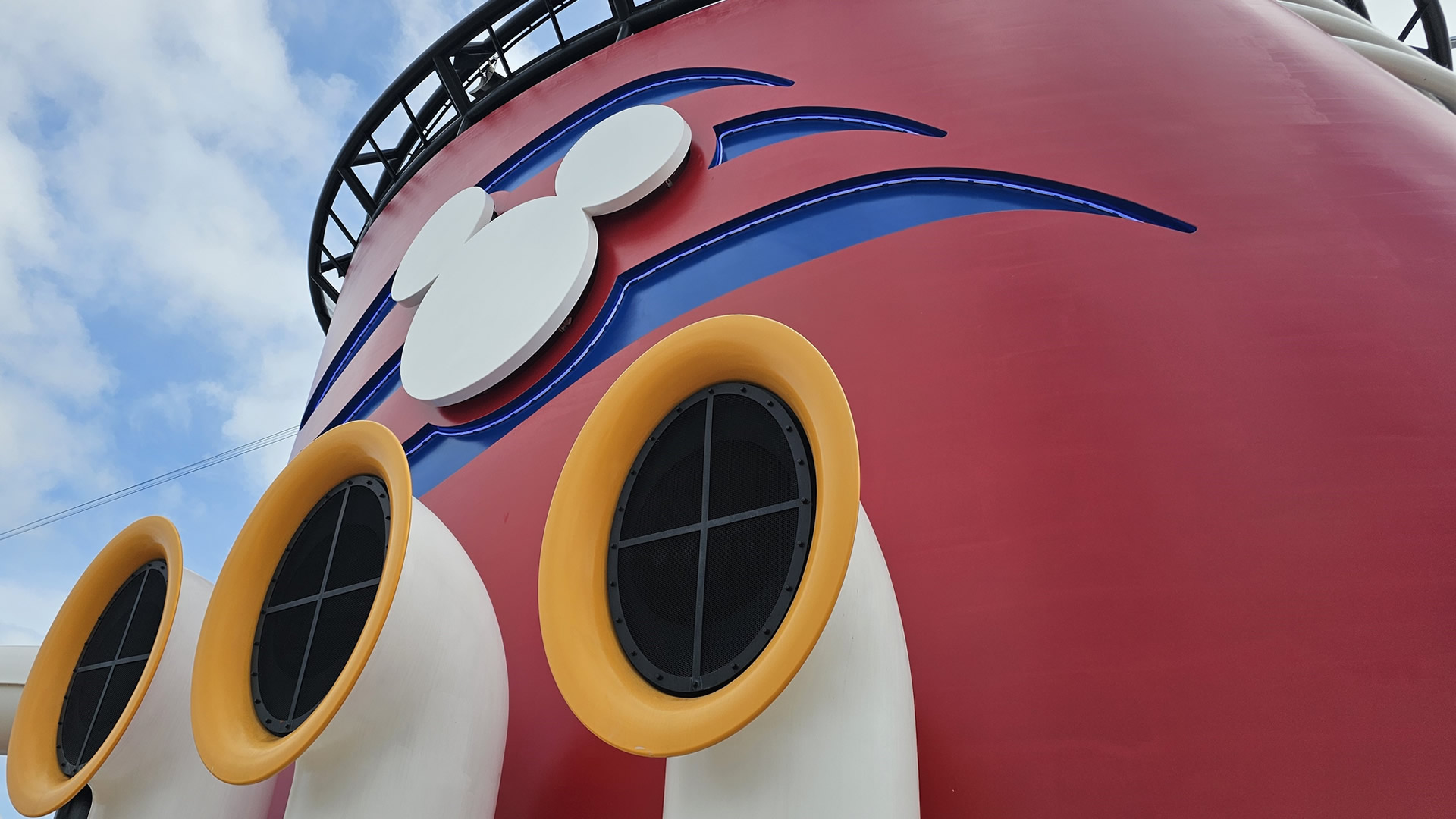 The red funnel of a Disney cruise ship is seen under a blue sky. The funnel features the blue and white Disney Cruise Line logo and yellow and white horn speakers.