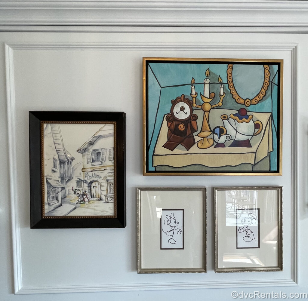 Framed art pieces depicting Mickey Mouse, Minnie Mouse, and characters from Beauty and the Beast are displayed in a One Bedroom Villa at Disney’s Riviera Resort. 3 of the pieces are drawings featuring Mickey and Minnie, and the largest is an abstract painting of Cogsworth, Lumiere, Mrs. Pott’s and Chip on a brown table with a blue background.