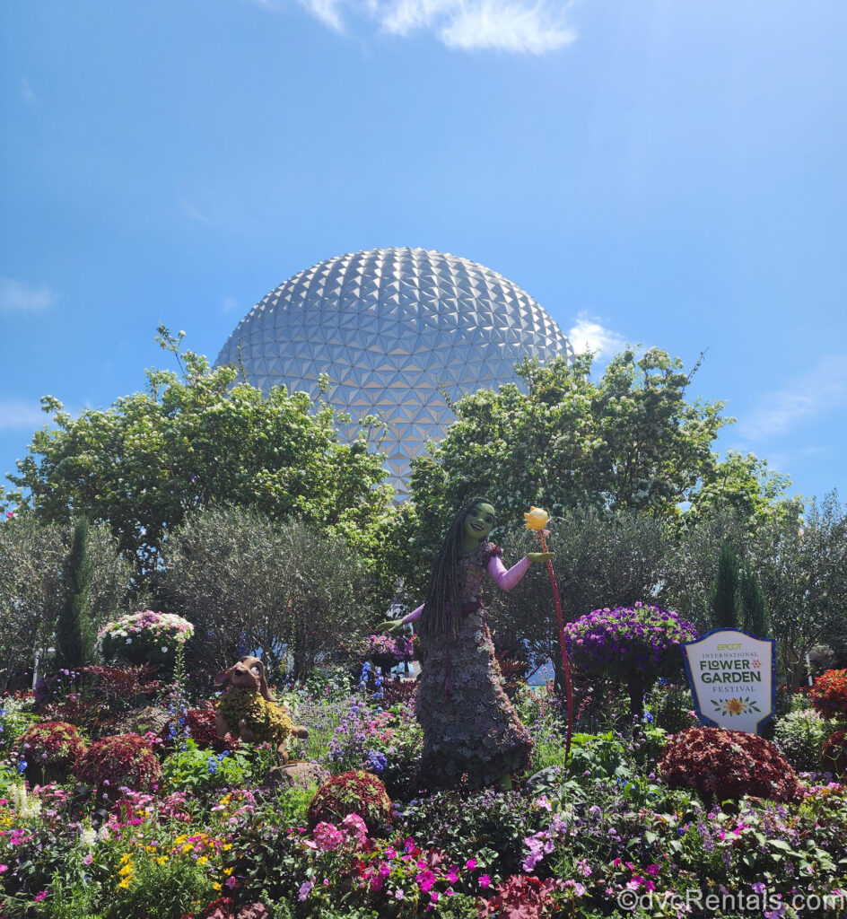 Mulitcolored flowers, and foliage topiaries of characters Asha, Valentino, and Star from the Disney movie Wish are seen in front of the Spaceship Earth ball. There is also a white sign reading “EPCOT International Flower & Garden Festival” in green lettering.