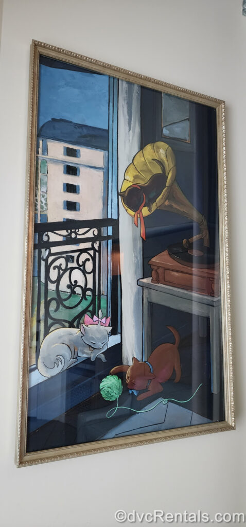 A painting featuring Marie, Toulouse, and Berlioz from the classic film The Aristocats shows the cats sleeping and playing around an old-fashioned wood and brass phonograph. The painting also depicts a hidden Mickey in the black balcony railing behind Marie and the grey resort exterior is seen in the background.