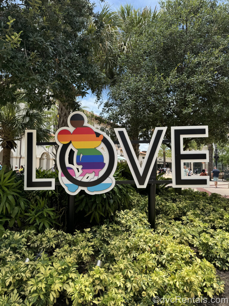 A sign reading the word “LOVE” in black and white lettering is displayed at Disney Springs. There is a rainbow-colored silhouette of Mickey Mouse poking through the center of the letter “O” in the sign to celebrate Pride.