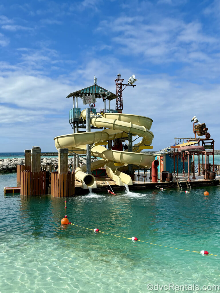 The twisting yellow tubes of the Pelican Plunge waterslides at Castaway Cay empty into the clear Caribbean water. The slides are on a large wooden platform along with a small waterplay area.