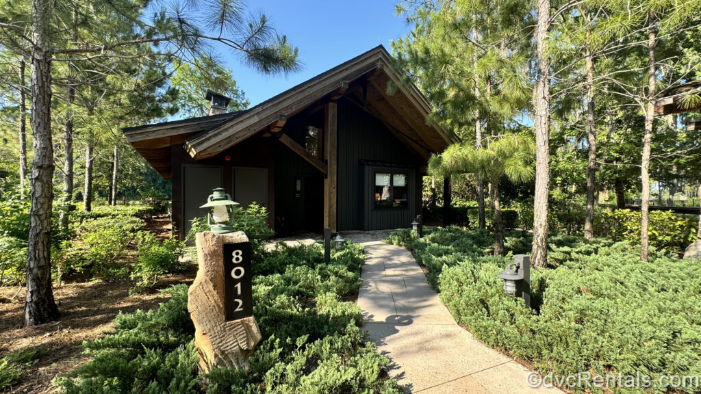 The green and brown exterior of a Cascade Cabin at the Copper Creek Villas & Cabins at Disney’s Wilderness Lodge. The cabin is surrounded by green foliage with a brown stone pathway leading up to it.