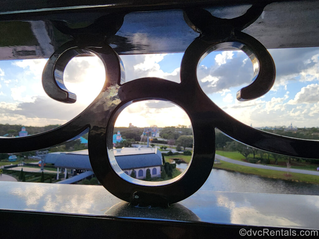 A black wrought-iron Mickey-head silhouette is seen in the balcony railings at Disney’s Riviera Resort. In the distance, the resort grounds including the Skyliner station and sunny blue sky can be seen.