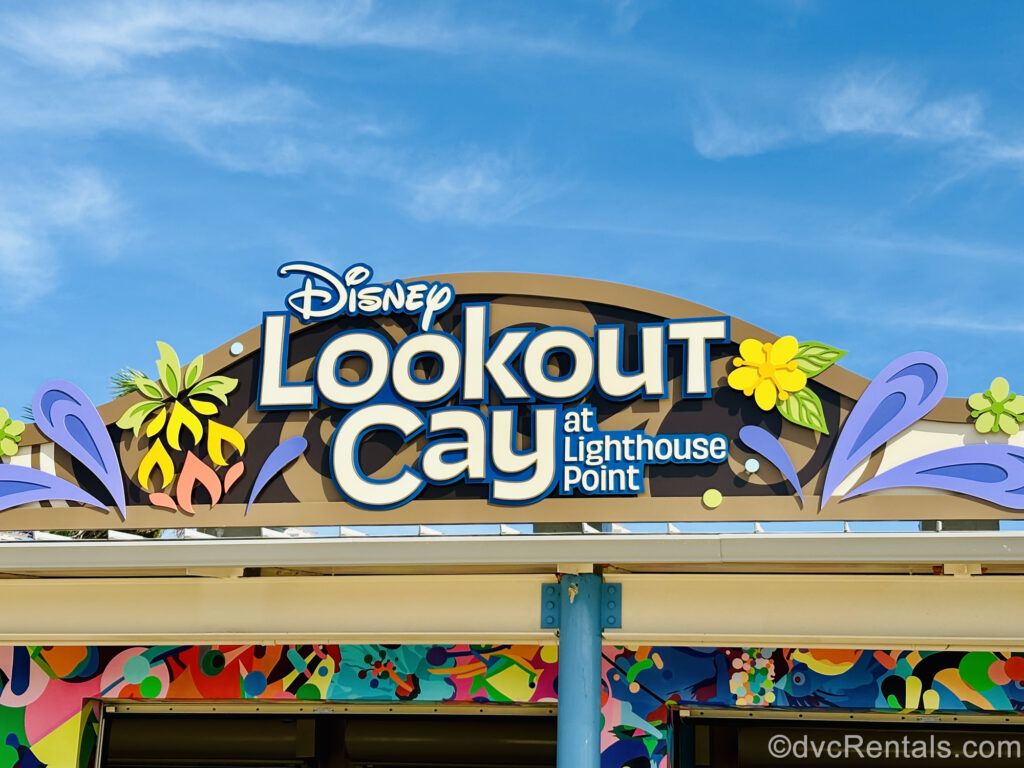 A sign reading “Disney Lookout Cay at Lighthouse Point” in white and blue lettering is seen under a blue sky. The sign is also decorated with vibrantly colored flowers and geometric patterns.