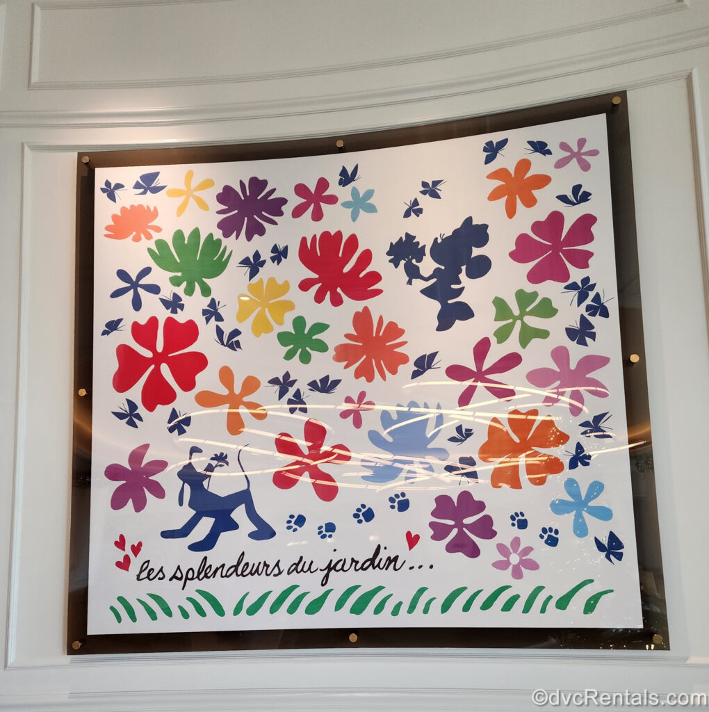 A colorful art piece displays navy blue drawings of Minnie Mouse and Pluto both holding flowers, with lots of multicolored flowers and butterflies around them, as well as some paw prints and a white background. At the bottom of the piece, the French phrase “les splendeurs du jardin” is written in black script.