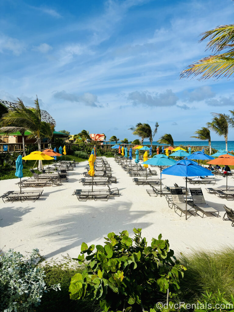 Lounge chairs on the white sand beach of Lookout Cay at Lighthouse Point. There are yellow, blue, and orange umbrellas amongst the loungers, and the curvy lines of building roofs can be seen in the background. In the distance there is turquoise water beyond the beach.