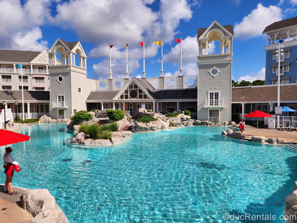 The Stormalong Bay pool area at Disney’s Beach Club Villas is seen under a sunny sky. The blue and grey resort buildings are seen in the background along with two lifeguards on either side of the pool.