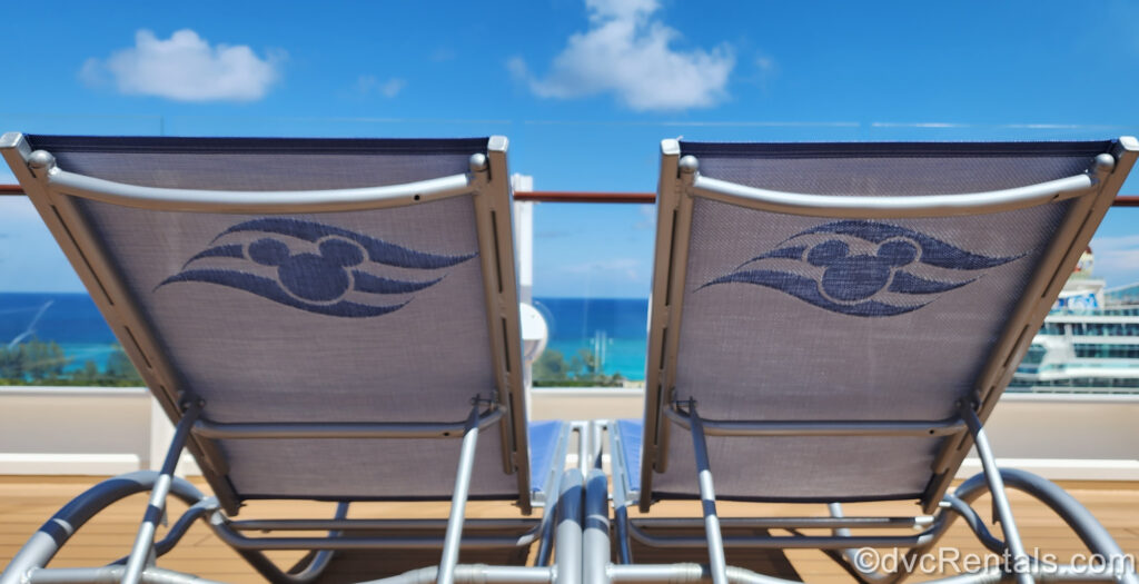 2 lounge chairs featuring the blue and white Disney Cruise Line logo with Mickey’s head in 3 waves sit in the sunshine looking out at the ocean on the wooden deck of a cruise ship.