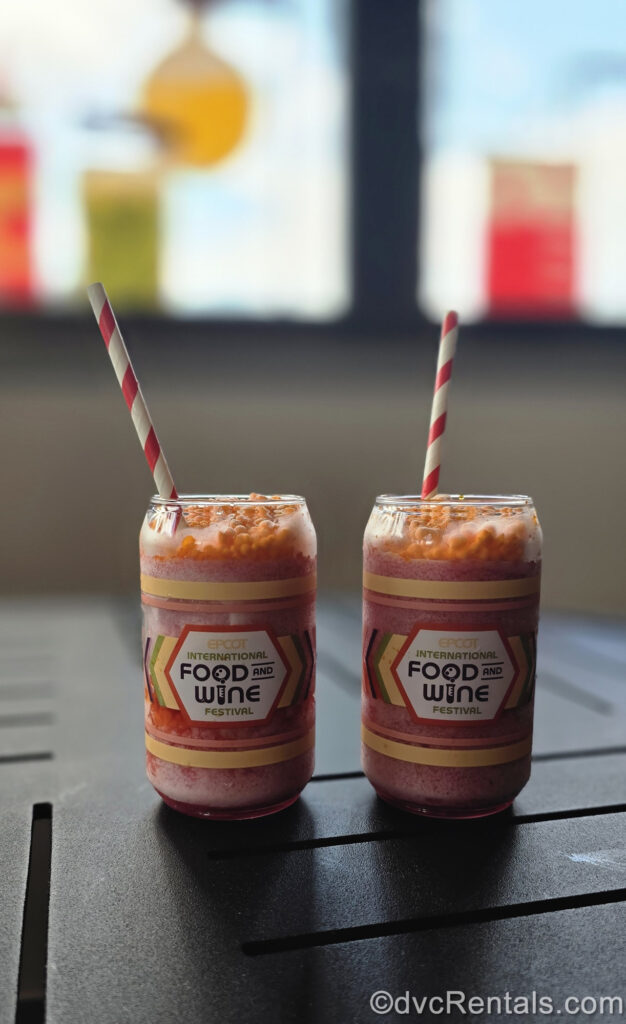 2 pink and orange beverages in glasses reading “EPCOT International Food and Wine Festival” in black and green writing are seen on a black table. Each drink has a red and white striped straw in it.