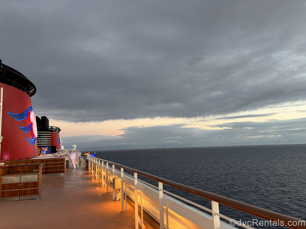 The red funnels and wooden decks of a Disney cruise ship are illuminated as the ship sails through the ocean which is visible on the right. The sky is cloudy with peeks of yellow and pink as the sunrise begins.