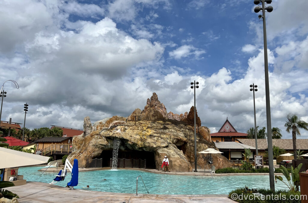 The Lava Pool at Disney’s Polynesian Villas & Bungalows. Water falls from the large brown stone structure made to resemble a volcano, as the brown and orange resort buildings are seen in the background.