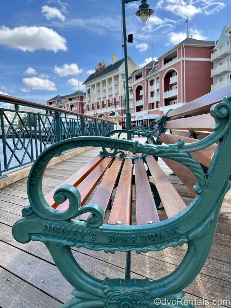 The green armrest of a wooden bench reading “Disney’s BoardWalk” is in the foreground while the pastel pink, yellow, and blue buildings and wooden planks of the boardwalk are seen in the background under a blue sky.