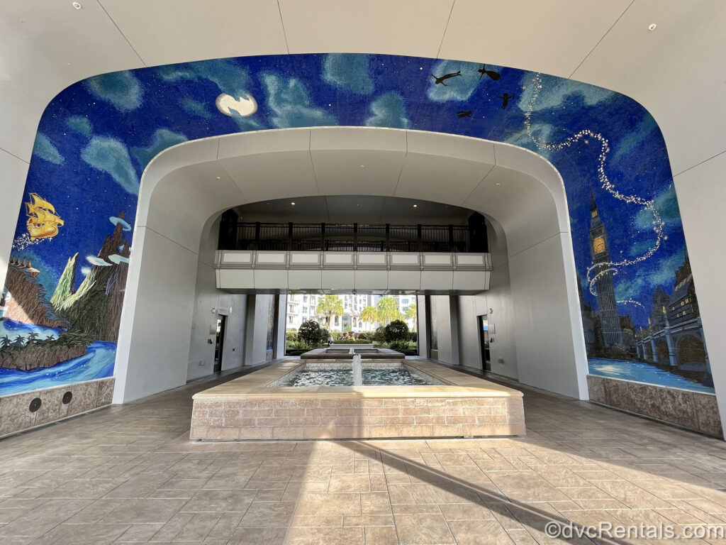 A large mosaic depicting Peter Pan and the Darling children flying through the night sky over the London skyline to Neverland covers the walls and ceiling over a fountain and walkway into Disney’s Riviera Resort.