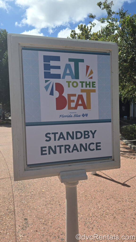 A white sign reading “Eat to the Beat Standby Entrance” in blue, green, orange, and red lettering is displayed at EPCOT during the International Food & Wine Festival.