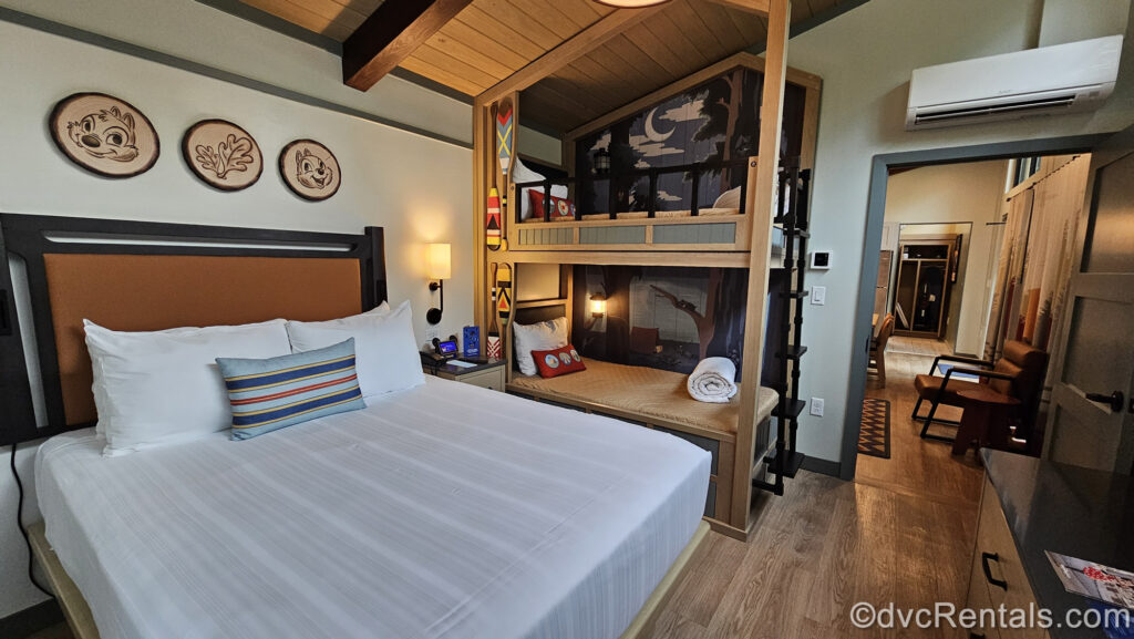 The bedroom of a One Bedroom Cabin at Disney’s Fort Wilderness Resort & Campground decorated in earthy tones. There is one king bed and bunk beds, with wooden artwork featuring Chip ‘N” Dale. Through the doorway, a chair in the living area and an open storage cabinet are also visible.