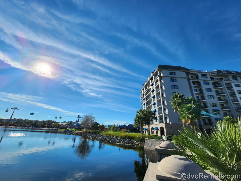 The Disney Skyliner is visible in the distance leading away from the station at Disney’s Riviera Resort. The grey and beige resort building sits next to water under a sunny, blue, sky.