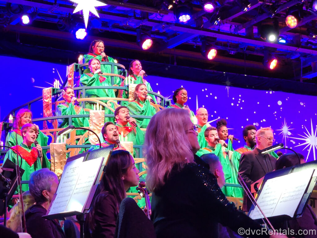 A choir dressed in green robes sings behind a live orchestra during Candlelight Processional at EPCOT. The stage is decorated with blue, red, green, and gold Christmas décor.