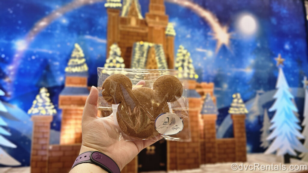 A hand holds a Mickey-shaped gingerbread cookie in front of the large Cinderella Castle gingerbread display at Disney’s Contemporary Resort.