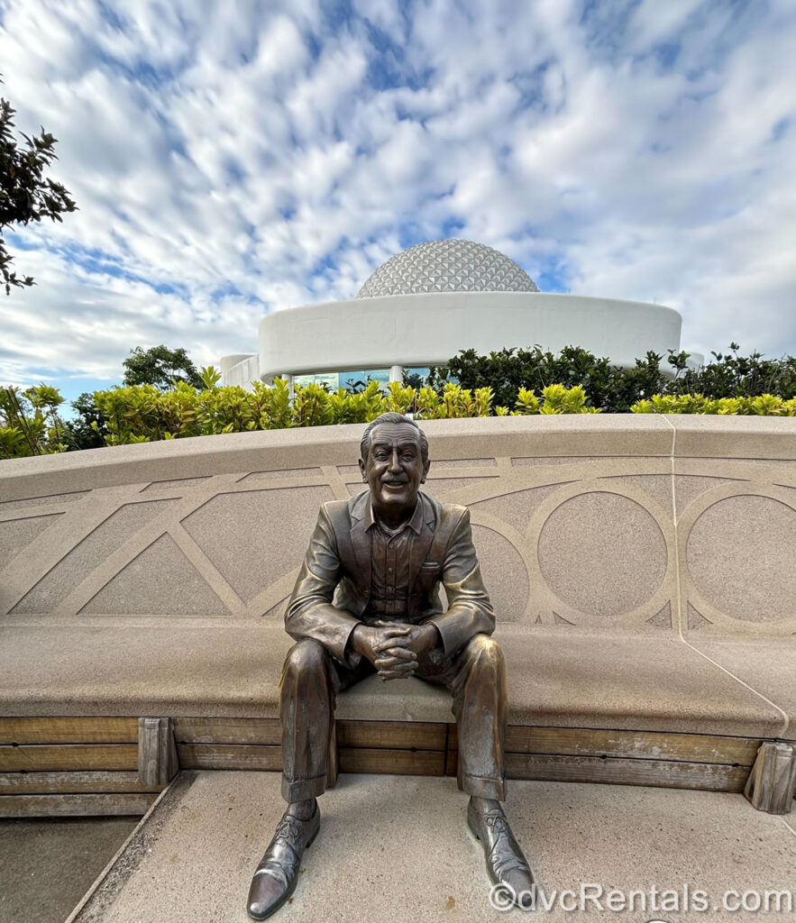 A bronze statue of Walt Disney sits on a stone bench at EPCOT. The Spaceship Earth ball is visible in the background along with a blue sky and green foliage.