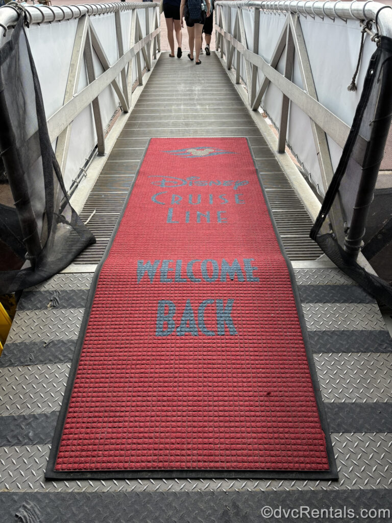 A red carpet reading “Disney Cruise Line, Welcome Back” in navy blue lettering is rolled out on the gangway ramp of a Disney cruise ship. Guests can be seen walking at the end of the ramp.