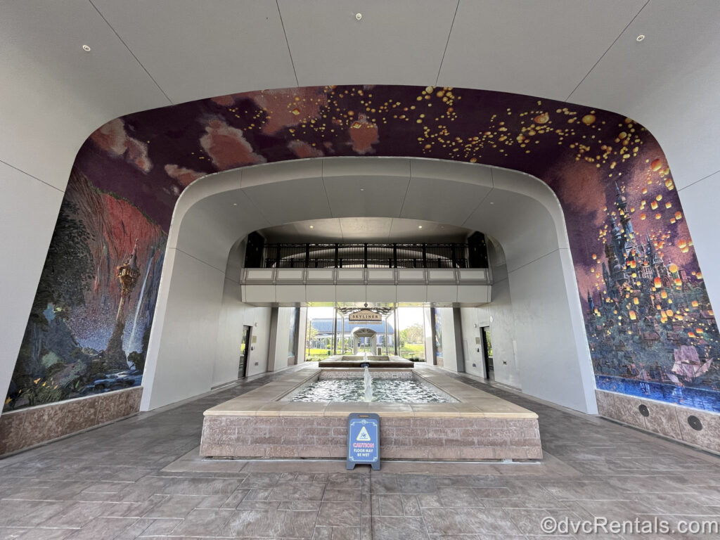 A large mosaic depicting Rapunzel’s tower, castle, and floating lanterns in a purple nighttime sky covers the walls and ceiling over a fountain and walkway into Disney’s Riviera Resort.