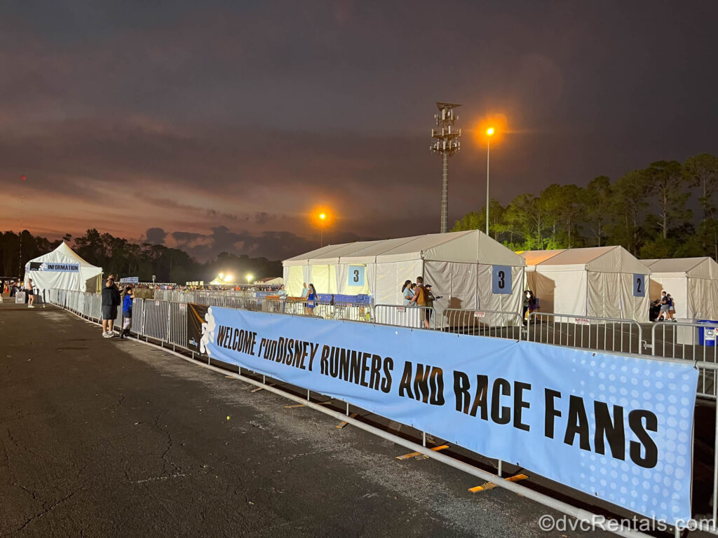 A blue banner reads “Welcome runDisney Runners and Race Fans” in black lettering. Behind the banner there are white, numbered tents, race participants, and the sun is just starting to rise in the darkened sky.