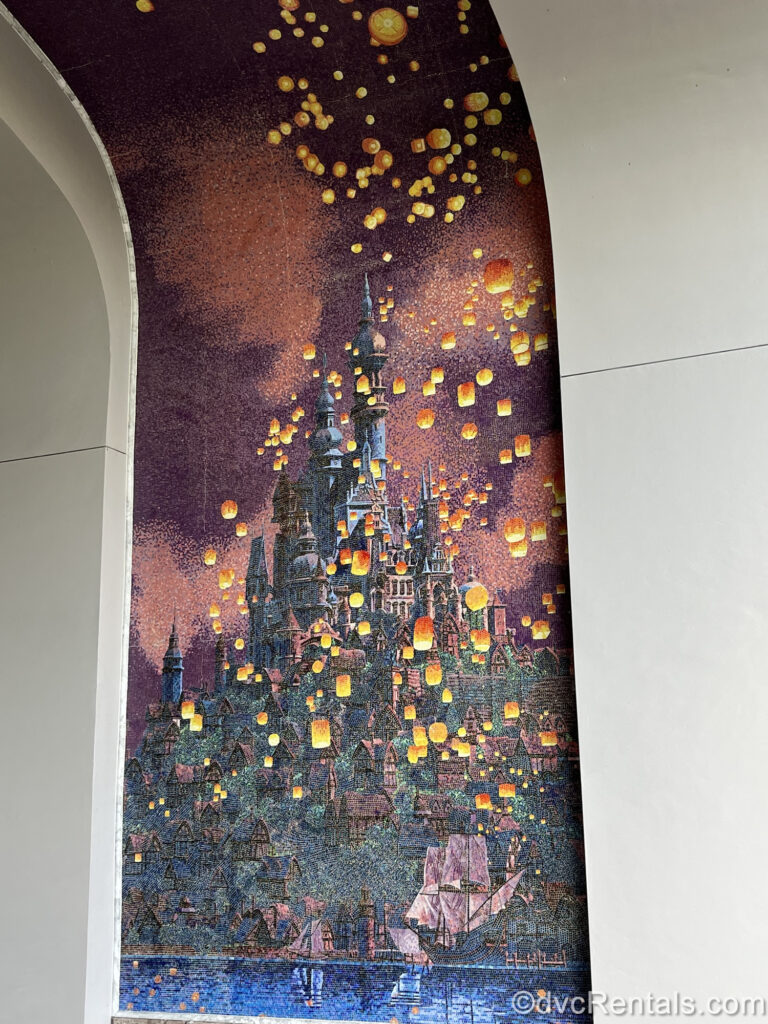 A mosaic of Rapunzel’s castle under a purple, nighttime sky is seen. There are numerous yellow and orange lanterns floating around and above the castle, and the small village is also depicted.