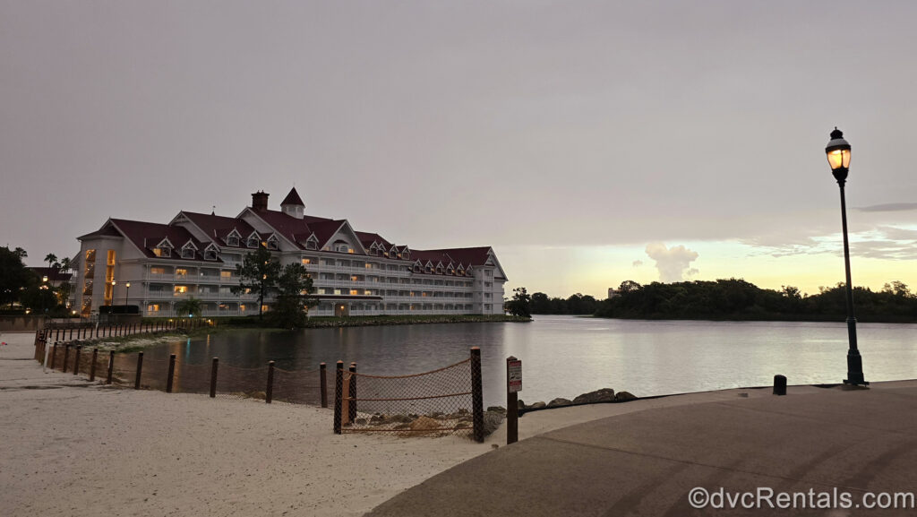 A red-roofed white building of Disney’s Grand Floridian Resort & Spa sits next to the water of the Seven Seas Lagoon as the sun sets. A white sand beach and lit lamppost are seen in front of the resort building.