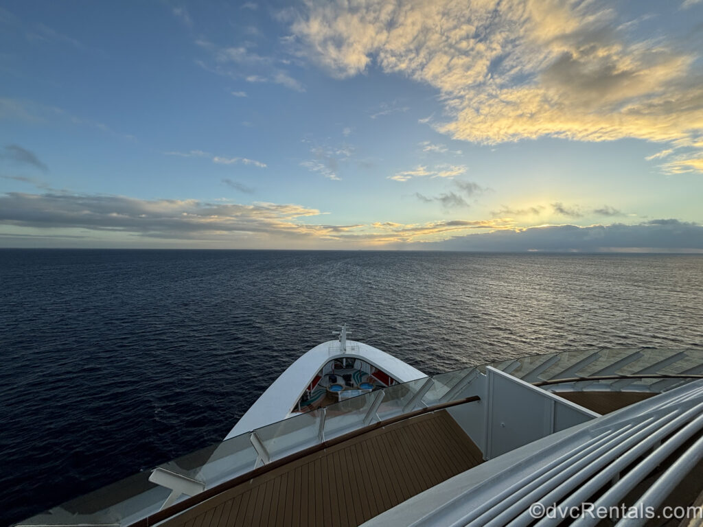 The view of the ocean at sunrise looking out from the white and wooden ship deck and railing. The sky is blue with white fluffy clouds illuminated by early morning golden sunlight.