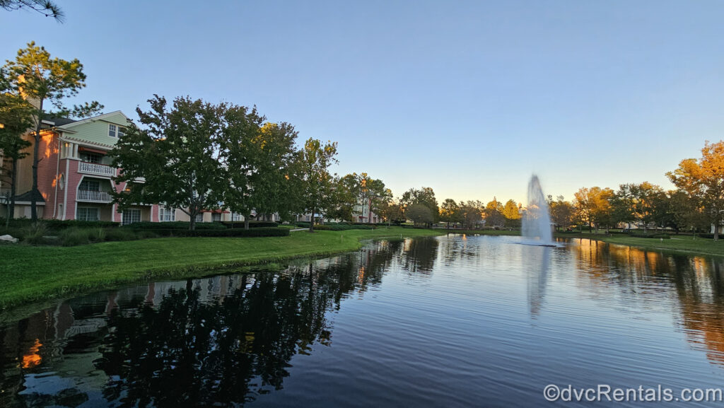 A fountain is seen in the water at Disney’s Saratoga Springs Resort & Spa. On the left side of the water the pastel pink and green resort buildings are seen, with sunshine shining on the trees around them.