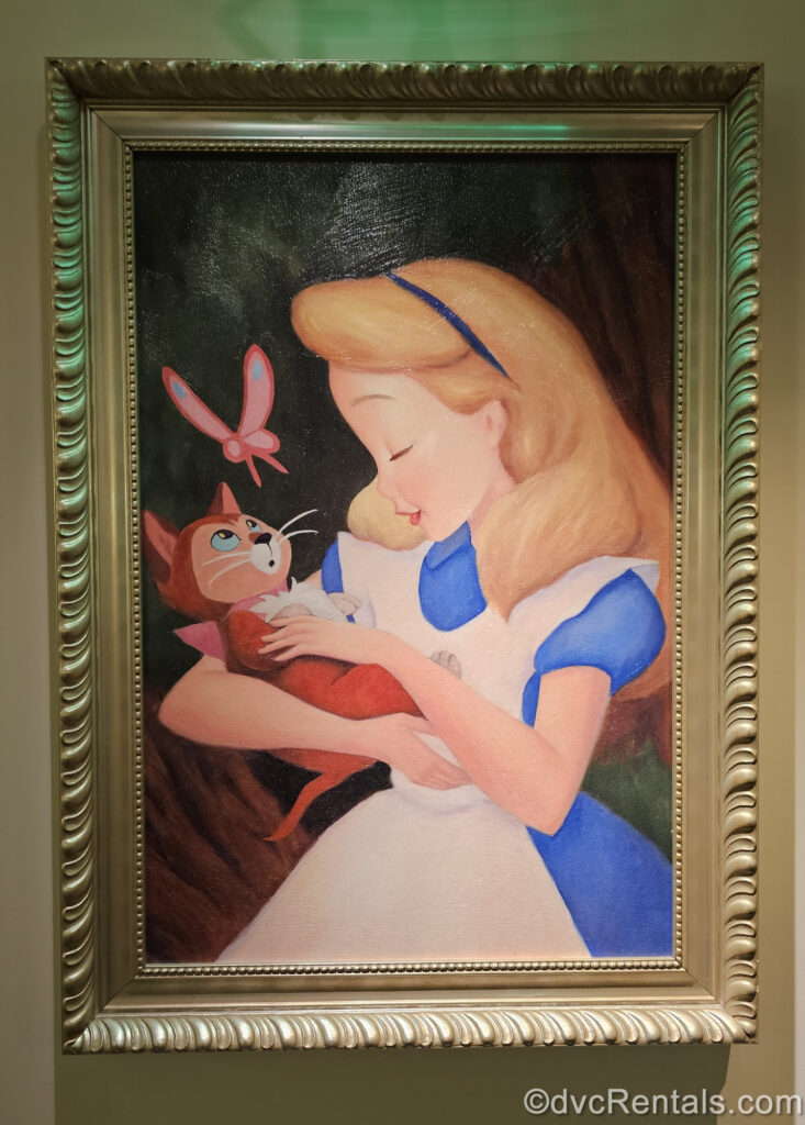 A painting of Alice in Wonderland in a gold frame. Alice wears her classic blue dress with white apron, and holds Dinah, her brown cat who is looking up at a pink butterfly.