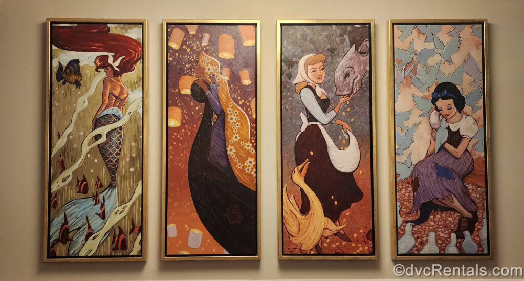 Four paintings, each of a Disney princess, are displayed in gold frames. From left to right the artworks feature Ariel as a mermaid with flounder in the sea, Rapunzel looking up at her lanterns, Cinderella feeding her horse and a goose, and Snow White with some blue birds.