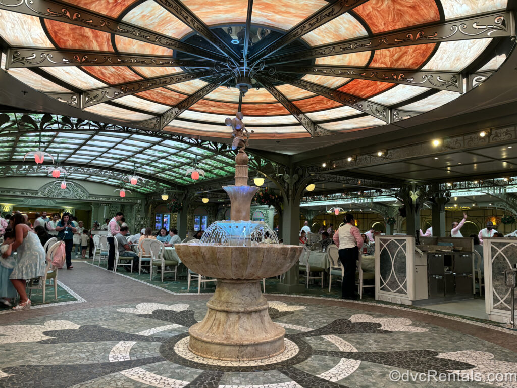 A beige stone fountain topped with a Mickey sculpture at the Enchanted Garden restaurant. Guests sit at white tables and the ceiling is designed to look like a green garden trellis.