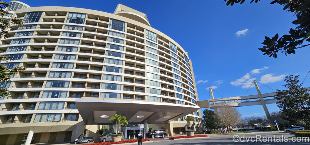 The beige building exterior of Bay Lake Tower at Disney’s Contemporary Resort is seen under a sunny sky. There are red flowers planted in front of the entry way and the Skyway Bridge leads to the Contemporary on the right.