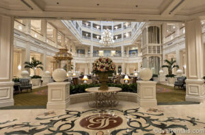 The main lobby of Disney’s Grand Floridian Resort & Spa. The resort’s monogram is seen in burgundy and white tiles on the lobby floor along with a large pink, red, and white flower display. The lobby is decorated mostly in neutral cream and green tones with Victorian influences.